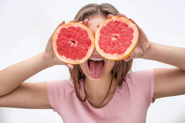 Die verspielte junge Frau bedeckt die Augen mit zwei Hälften Grapefruit. Musterschüler zeigen Zunge. isoliert auf grauem Hintergrund. — Stockfoto