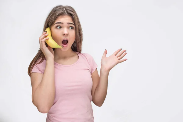 Mulher jovem surpreendente segurar banana em vez de telefone. Ela fala e acena com a mão. Isolado sobre fundo cinzento . — Fotografia de Stock