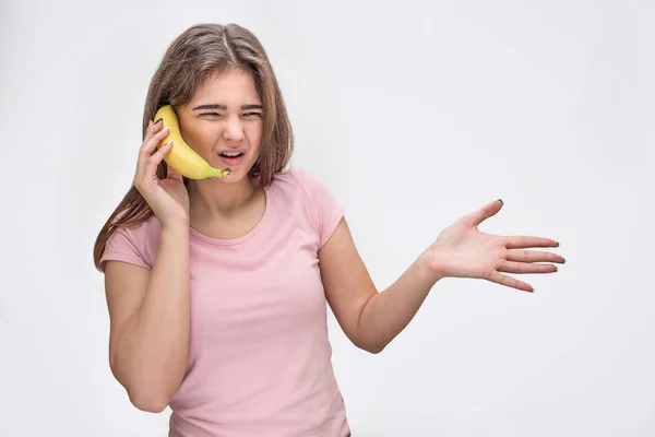 Отвратительная молодая женщина держит банан вместо телефона. Она говорит и показывает ладонь. Изолированный на сером фоне . — стоковое фото