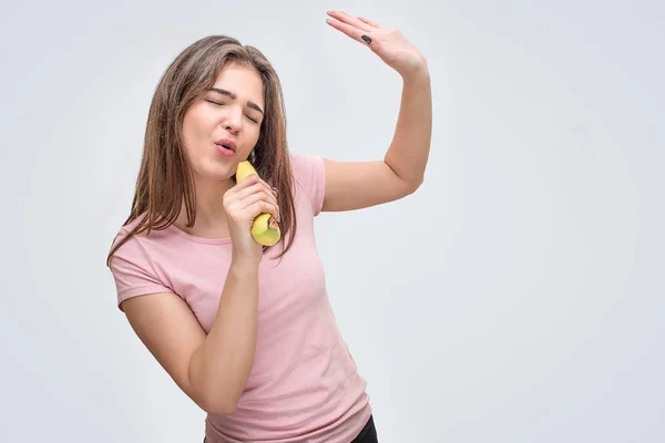 Веселая молодая женщина держит банан и поет в него. Она притворяется, что это микрофон. Наслаждайся. Изолированный на сером фоне . — стоковое фото
