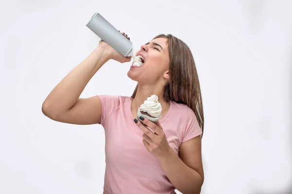 Фото молодой женщины, заливающей сливки в рот. Она держит торт в другой руке. Изолированный на сером фоне . — стоковое фото