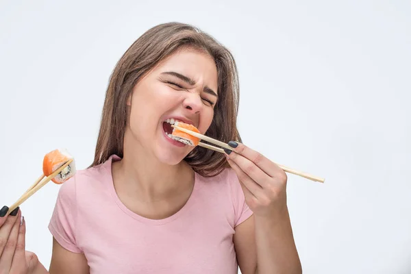 Молода жінка їсть шматочки суші. Вона кусає шматочок. Модель тримає їх за допомогою паличок. Ізольовані на сірому фоні . — стокове фото