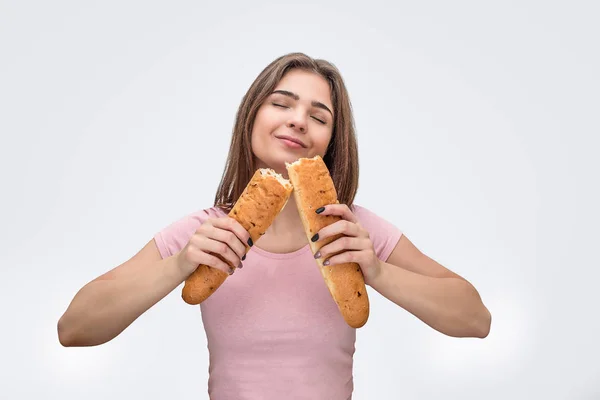 Delightful young woman smell fresh baguette. She hold both piees in hands. Model keep eyes closed. Isolated on grey background. — Stock Photo, Image
