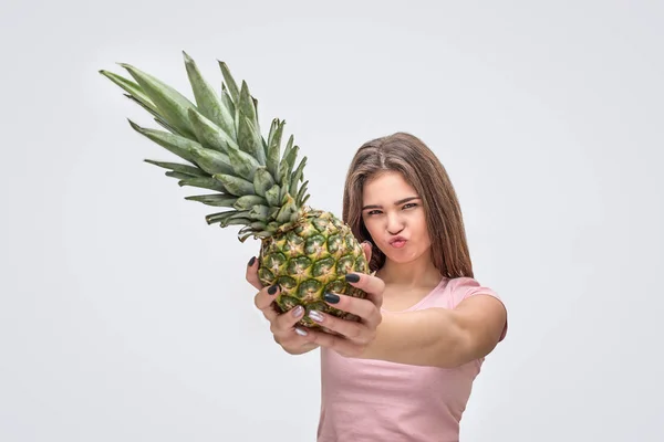 Серьёзная молодая женщина показывает ананас с рук. Она смотрит в камеру. Изолированный на сером фоне . — стоковое фото