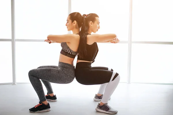 Dos mujeres jóvenes descuidadas ejerciendo en la sala de fitness. Mantienen las manos cruzadas en el área del pecho y se tocan entre sí. Las modelos se sientan en pose de escuadrón . — Foto de Stock
