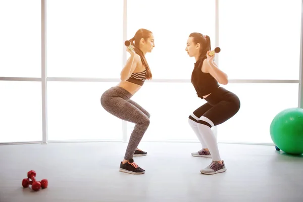 Zwei sexy junge Frauen beim Workout im Fitnessraum. Sie halten die Langhantel auf Schultern und in die Hocke. Modelle schauen einander an. — Stockfoto