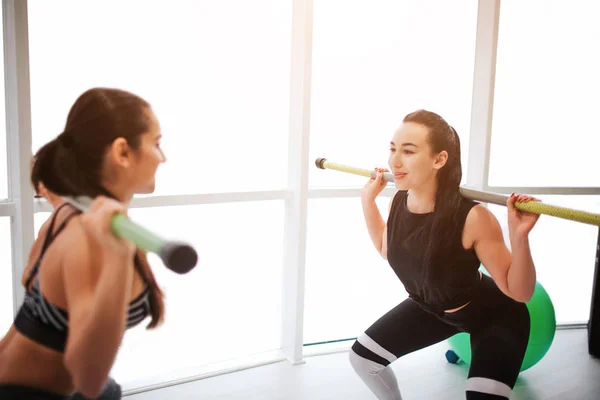 Vrolijke jonge vrouw samen oefenen. Ze houden de halter op de schouders en squadding. Model blik op elkaar en glimlach. Ze trainen in fintess kamer. — Stockfoto