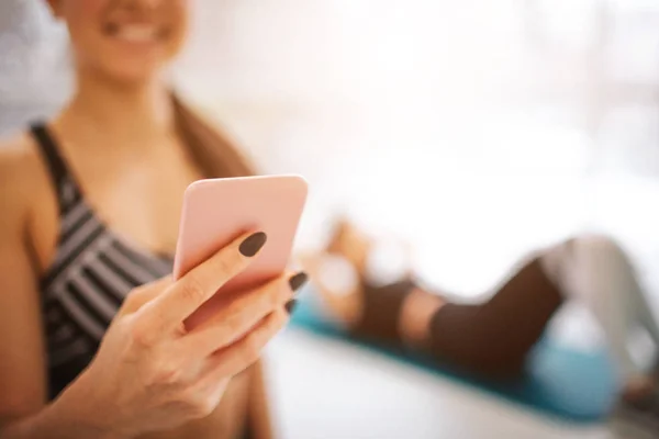 Geknipte weergave van vrolijke jonge vrouw houd de telefoon in de hand en glimlach. Ze staan in de fitnessruimte en kijken naar apparaat. Haar vriend doen abs oefening op matras. — Stockfoto