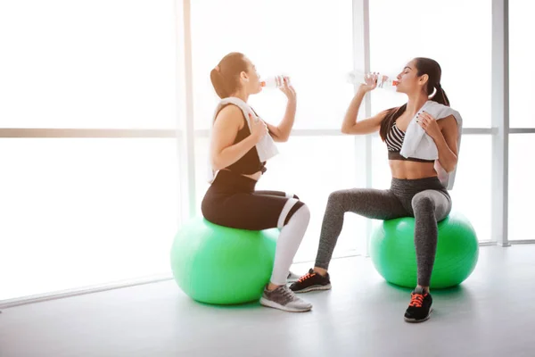 Due belle e magre giovani donne si siedono su palline verdi in sala fitness. Bevono acqua dalle bottiglie. I modelli hanno riposo dopo l'allenamento . — Foto Stock