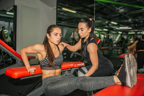 Sexy young woman working hard in gym. She exercise at bench. Model does abs. Asian female trainer help her.