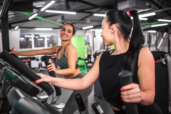 Mulheres jovens positivas e simpáticas olham umas para as outras e sorriem. Eles trabalham no ginásio em simulador cross-trainer . — Fotografia de Stock