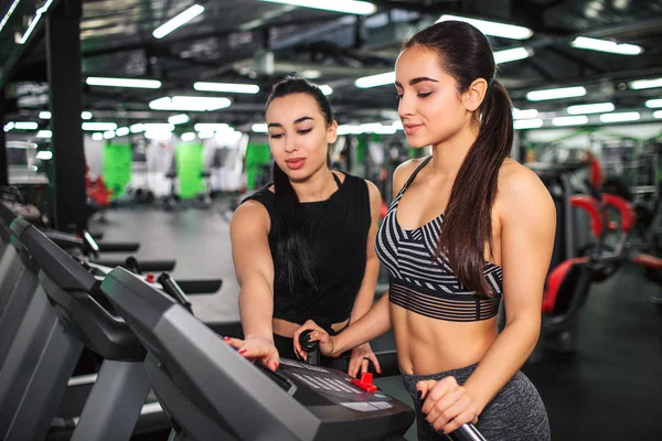 Mooie jonge vrouw staan op de atletiekbaan en kijk naar het deelvenster. Aziatische vrouwelijke trainer helpen haar aanpassen van de instellingen van de computer. Ze zijn in sportschool. — Stockfoto