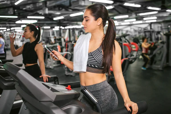 Sexy mladá žena stojí na běžecké dráhy a telefon. Dívá se na to a úsměv. Asijské mladé ženy stojí na stroji příliš a pít vodu. — Stock fotografie