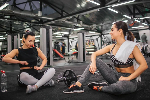 Bild von zwei jungen Frauen, die auf dem Boden in der Turnhalle sitzen. asiatische Modell Krawatte Knoten auf Hemd und Blick nach unten. junge Europäerin schaut ihn an und hält weißes Handtuch auf der Schulter. — Stockfoto