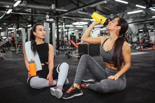 Zwei junge Sportlerinnen sitzen in der Turnhalle am Boden. asiatische Modell aussehen, wie ihr Freund Wasser aus gelben Flasche trinkt. sie hält orangefarbenes und weißes Handtuch. — Stockfoto