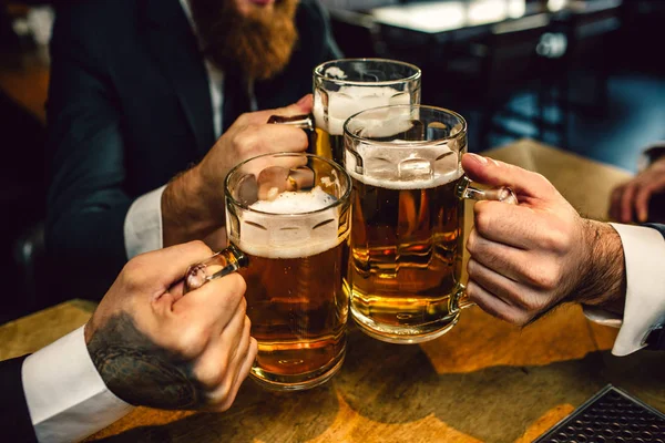 Drei Männer in Anzügen halten Bierkrüge zusammen. sie sitzen am Tisch. — Stockfoto