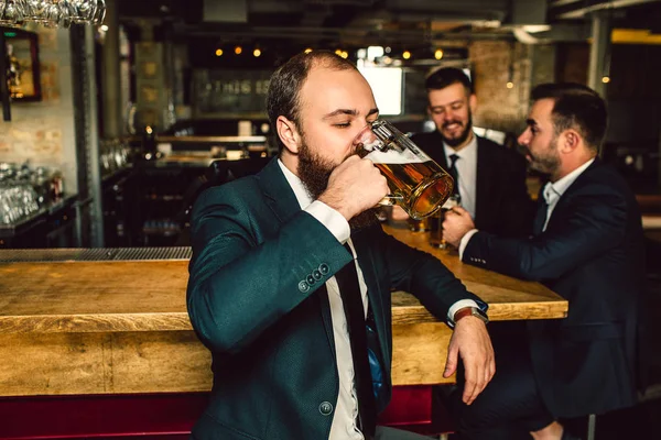 Fiatal férfi öltöny állvány sáv- és sört inni. Két férfi álljon a háta mögött. Ők beszélnek. — Stock Fotó