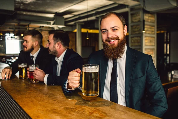 Pozitif ve mutlu genç işadamı oturup bar sayacı. Kamerada gülümsüyor. Adam tutun bira bardağı. Diğer iki ofis çalışanları arkasında oturmak. — Stok fotoğraf