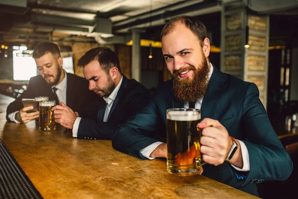 Hombre barbudo alegre y positivo en traje mirada en la cámara. Sonríe y sostiene la taza de cerveza. Otros dos jóvenes se sientan detrás . —  Fotos de Stock