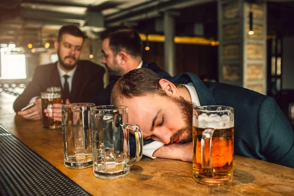 Tired young man in suit sleep on bar counter. He is drunk. there are two empty mugs and one full with beer. Other two young men sit behind. — Stock Photo, Image