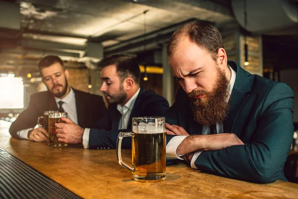 Uykulu genç adam bar counter uyku. Bira bardağı vardır. Diğer iki adam arkasında oturup konuşmak. Onlar-si olmak iki bira kupa. — Stok fotoğraf