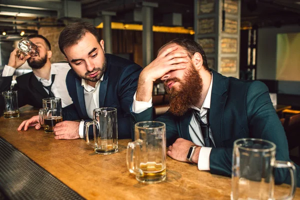 Young bearded man sit in front and sleep. He is drunk. Second young man look at him. Third drink beer. — Stock Photo, Image