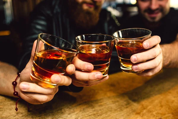 Skåret billede af tre skæggede unge mænd, der holder glas med rom sammen. De smiler. Folk sidder i bar . - Stock-foto