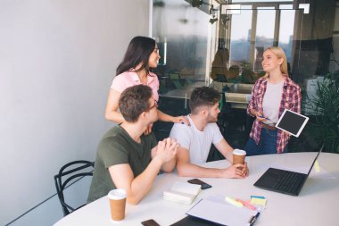 Ciddi genç adam masada oturup dizüstü ekrana bak. Esmer ve blone genç kadın onların arkasında dur ve gülümse. Bir odada beraberler.