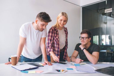 Neşeli genç kadın ve adam sağ gülümse ve aşağıya bak. Malzemeler ile tabloda işaret eder. Beyaz tişörtlü ciddi genç adam aşağı bak. Coworking içinde çalıştıkları.