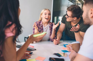 Neşeli sarışın genç kadın gülümse ve dört gözle bekliyorum. O el salla. Esmer kadın ve iki genç erkek ona bak. Dinliyorlar. İnsanların birlikte coworking içinde çalışma.