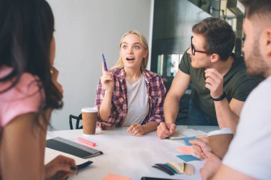 Neşeli sarışın genç kadın gülümseme ve sizi fikir bekliyoruz. O el salla. Esmer kadın ve iki genç erkek ona bak. Dinliyorlar. İnsanların birlikte coworking içinde çalışma.