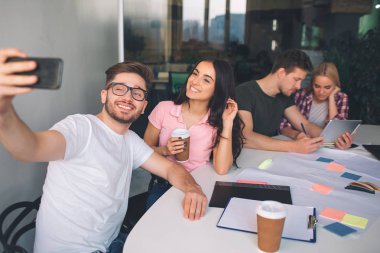 Genç adam tutarak resmi telefon ve selfie güzel esmer ile alın. Poz ve gülümse. Başka bir çift onların arkasında otur ve birlikte çalışır. Onlar beyaz odada oturup.