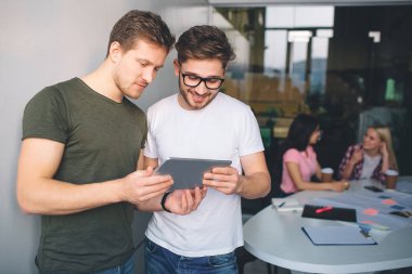 İki genç ve güçlü genç adam yan yana ve tablet vasıl bak. Merak ediyorlar. Genç kadın arkalarındaki masada oturup konuşmak. Bir beyaz odada insanlar.