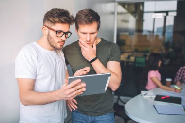 Ciddi ve yoğun genç erkek yan yana ve tablet vasıl bak. Düşünüyorlar. Genç kadınlar birlikte tablo ve iş onların arkasında oturmak. Bir beyaz odada insanlar.