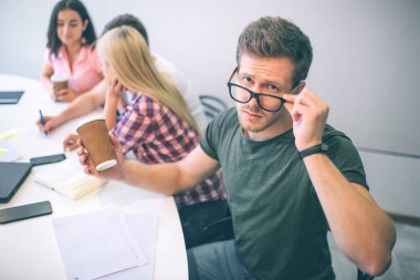 Ciddi genç adam kameraya bak. O gözlük elini tut. Kahve başka bir koludur. Arkadaşlarıyla onun arkasında otur ve birlikte çalışma.