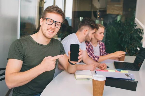 Bonito joven en gafas apuntan en hpone en las manos. Él mira en la cámara. Otros dos jóvenes trabajan juntos en un ordenador portátil . — Foto de Stock