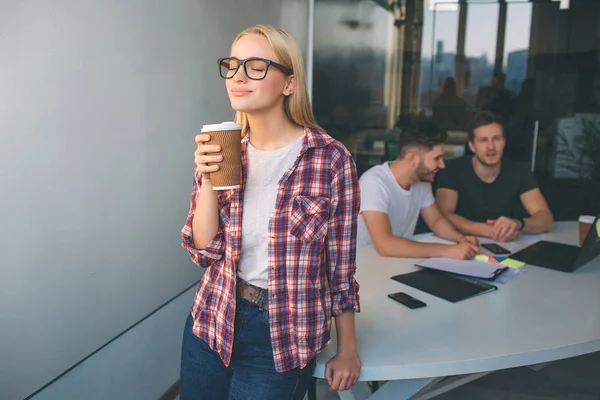 Smarta unga blonda kvinnan bära glasögon. Hon står och håller kopp kaffe. Hon enjoyes. Två unga män sitter bakom henne och jobbar. De pratar sinsemellan. — Stockfoto