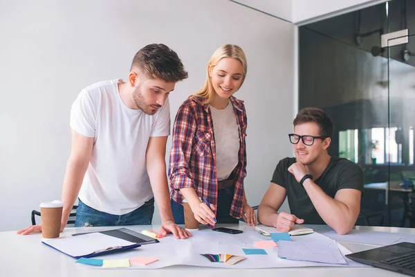 Joyeux jeune femme et l'homme sur le sourire droit et regarder vers le bas. Elle pointe sur la table avec des matériaux. Un jeune homme sérieux en chemise blanche regarde vers le bas. Ils travaillent ensemble dans le coworking . — Photo