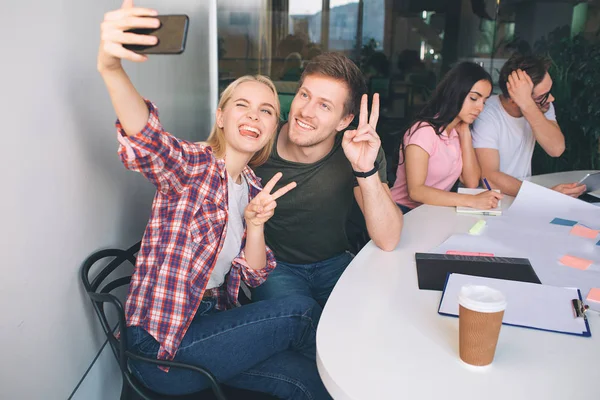 幸せな若いカップルが selfie の話。彼らは一緒に座るし、携帯電話のカメラを見ています。人々 を部分シンボル。別のカップルそれらの後ろに座るし、テーブルで働く. — ストック写真