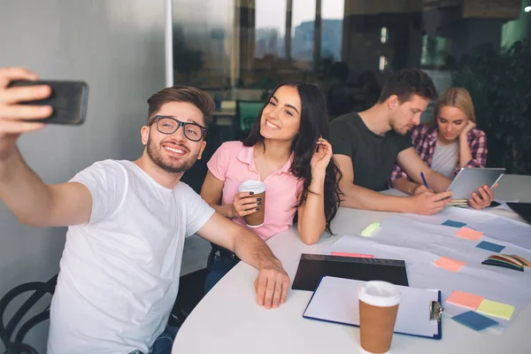 Foto eines jungen Mannes, der sein Handy hält und ein Selfie mit der schönen Brünetten macht. Sie posieren und lächeln. Ein weiteres Paar sitzt hinter ihnen und arbeitet zusammen. sie sitzen im weißen Raum. — Stockfoto