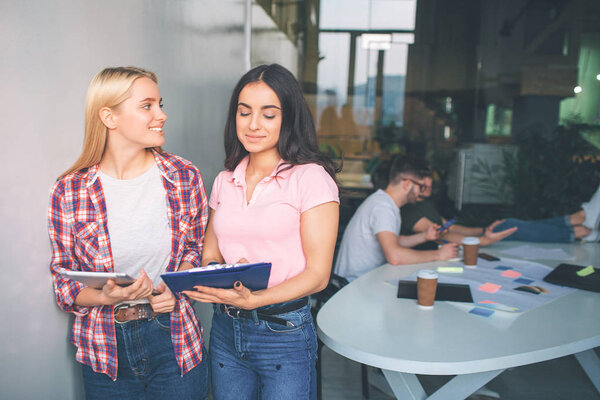 Nice and positive young women stand together in white room. Blonde model look at brunette. Second one look at tablet. Two men sit behin them and work.