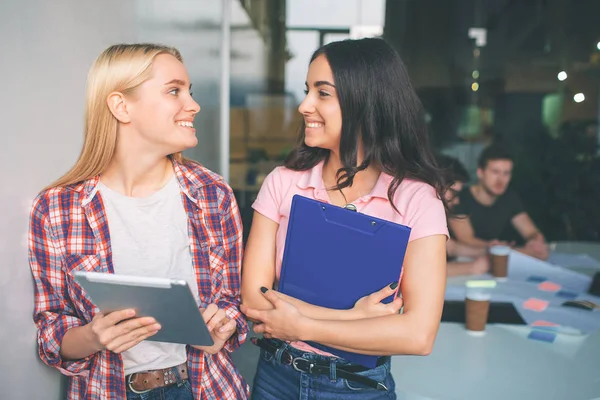 Bild der jungen blonden und brünetten Models stehen zusammen und lächeln. Sie sehen glücklich aus. Junge Frauen halten elektronische und Plastiktabletten in den Händen. Dahinter arbeiten zwei junge Männer. — Stockfoto