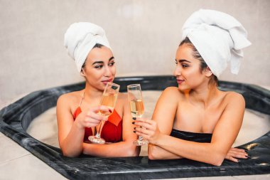 İnce ve seksi genç kadın jakuzide oturun ve birbirlerine bakıyorlar. Modelleri Gözlük şampanya ile dokunun. Vücut beyaz havlu ile sarılır.