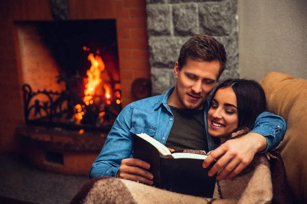 Positive and cheerful couple sit together. Young woman covered with blanket. Guy read book for her. They sit near fireplace. — Stock Photo, Image