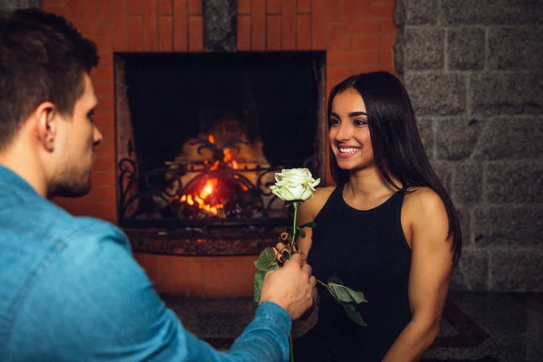 Jovem alegre sorrir para o namorado. Ela olha para ele. O tipo deu-lhe uma rosa branca. Sentam-se na lareira . — Fotografia de Stock