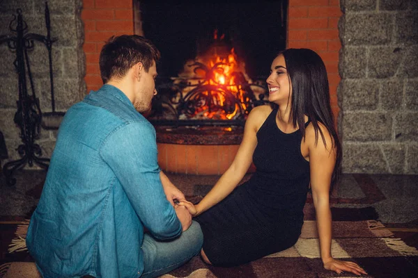 Um jovem sério senta-se no chão com a namorada na lareira. Ela olha para ele e sorri. O tipo segurou-lhe a mão. Eles namoro . — Fotografia de Stock