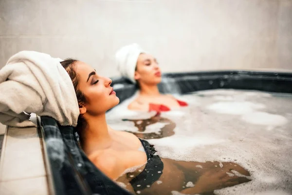 Obraz dwa zrelaksowany młodych kobiet chłodzenie z zamkniętymi oczami. Europejski model siedzieć z przodu. Azjatyckie, jest na plecach. Noszą stroje kąpielowe i ręczniki wokół włosów. Woda jest pełna piany. — Zdjęcie stockowe