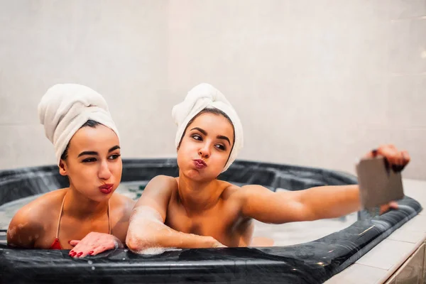 Belos modelos jovens sentar juntos em jacuzzi. Mulher europeia segurar o telefone e tirar selfie. Modelo asiático pose. Seu cabelo é envolto com toalhas brancas . — Fotografia de Stock