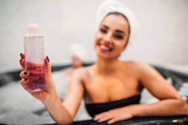 Çekici genç kadın duş jeli el ve gülümseme şişe tutun. Doğrudan kameraya bak. Siyah mayo ve saç etrafında beyaz havlu modelleri giymek