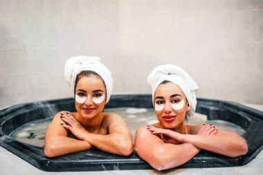 Neşeli genç modelleri jakuzide oturun. Su ve köpük dolu. Genç kadın doğrudan kameraya bak. Onlar göz altında gözleri ve havlu saç çevresinde var.